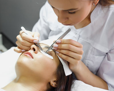 Aesthetician with tweezers applying eyelash extensions to a woman laying down, with her eyes closed.