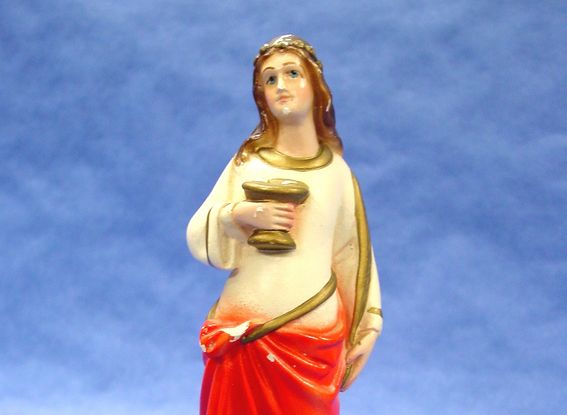 A small statue of a woman wearing a robe on a blue background. She has light skin and long brown hair, she wears a white and red robe, and holds a gold palm frond in one hand and a gold platter with two eyeballs on it in the other.