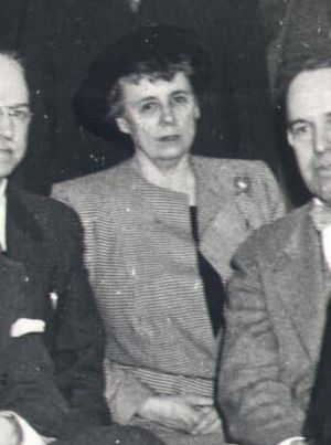A zoomed-in black and white photograph of a woman sitting in a group of men. She is an older white woman wearing a houndstooth coat and a round black hat over her short hair. She sits between two older white men wearing suits, and she is making eye contact with the camera.