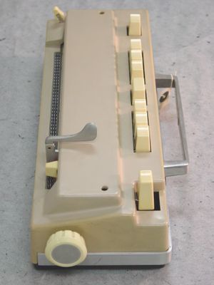 A typing machine made from beige plastic. The machine has nine tall white buttons for typing and a white knob on the side. One side has a silver-colored switch and the other side has a silver-colored handle.