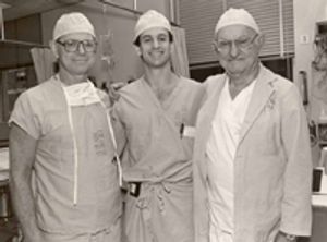 A black and white photo of three doctors wearing scrubs have their arms around one another. They are all white men. The man on the left is middle aged and is wearing dark framed eyeglass, a surgical mask is hanging on his chest. The middle man is the youngest, and he has dark hair and wears a beeper on his waist. The man on the right is the oldest, and he wears eyeglasses and a lab coat over his scrubs. They are all smiling jovially.