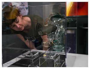 A young white woman with dark hair looks into a museum artifact case. The case has two pairs of eyeglasses and a clear, glass headform that is wearing a black set of magnifying lenses. All of the objects sit on clear plastic risers, and an orange TV screen can be seen in the background.