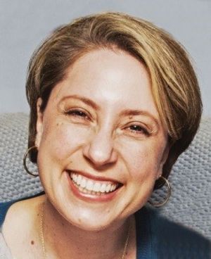 A woman smiles at the camera. She is a white woman with short cropped brown hair, and she wears lipstick and hoop earrings.