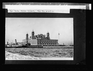 A black and white photograph of a brick building is taped to a black sheet of paper. The building is made of brick and has four turrets, and there is an American flag on a flagpole in front. A black steamship with a funnel is pulled up next to the building. The building appears to be surrounded by water. Text across the bottom of the photograph reads: New York. Ellis Island. Neg. No.