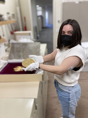 A young woman wearing a white sweater, blue jeans, white cotton gloves, an insulin pump, and a black medical mask holds a large gold medal. She has light skin and short black hair, parted in the middle. She stands in a beige room and holds the medal in her outstretched hands.