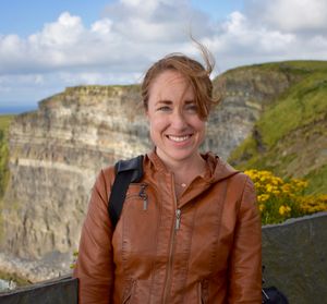A young white woman with red hair stands outside in front of a cliff. She is wearing a brown leather jacket and her hair is being blown by the wind. There are yellow flowers over her right shoulder and the cliff face is grey with green grass on the top. There are white clouds in the blue sky.