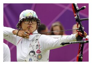 A photograph of a young Korean woman firing a compound bow and arrow. She is wearing a white collared shirt and a white bucket hat with plaid lining. She also wears large, black framed glasses and keeps both of her eyes open. She is drawing back the string of the bow, and holding it against her lips. 