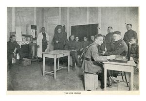 A black and white photograph of a group of soldiers. One soldier is performing an exam for eyeglasses on another solider. The man performing the exam is wearing a uniform and black leather boots, and he is pulling lenses from a large case on a table. The man receiving the exam is wearing a large military overcoat and has a pair of glasses with empty frames to hold test lenses. Seven other men in military uniforms sit behind them.