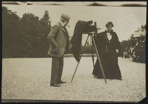 A sepia-toned photograph of a man and a woman standing near a camera on a tripod. He is an older, white man with white hair wearing a tweet suit and cap, and he stands with a pipe in his mouth and his hands in his jacket pockets. She is a white woman with dark hair wearing a long black black dress with a black cape, and her hair is pulled up on the top of her head. The camera is a large, early 1900s-style camera with an accordion-like structure in the middle and a large black cloth for the photographer to duck underneath.