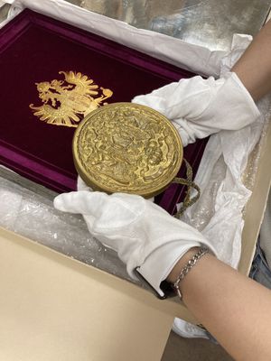 A person wearing white cotton gloves holds a large gold medal over a purple velvet book cover with a gold imperial eagle symbol. The book sits in a beige box full of white tissue paper.