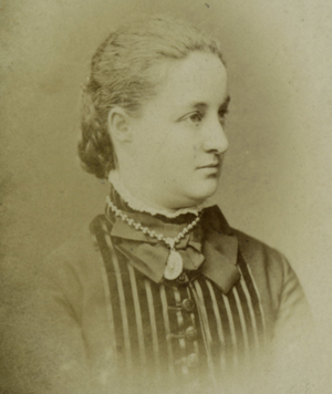 A sepia-toned photography of a woman. The photo is from her shoulders up, and she is posed looking off to the right of the camera. She is a young, white woman with her hair pulled in a bun at the base of her neck. She is wearing a dark, high collared, 19th-century style dress and a beaded necklace with a small seashell pendant.