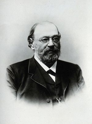 A black and white portrait of an older white man wearing a dark black suit. He has dark curly hair and a large forehead, and he wears a beard and small circular eyeglasses.