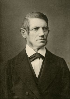 A black and white photograph of an older white man wearing a black suit. He has a slick, 19th century hairstyle, and wears a black ascot tie with his suit. He is looking to the right of the camera with a stern expression.