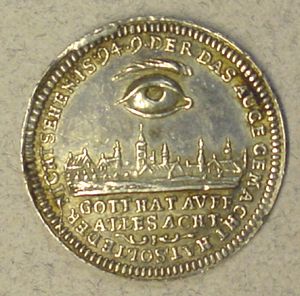 A small silver metal coin sits on a gray background. The coin depicts a human eye and eyebrow floating over a nineteenth-century-style city scape. The German text across the bottom of the coin reads: GOTT HAT AUFF ALLES ACHT.