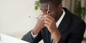A man in a business suit rubs his eyes while holding his glasses and sitting in front of a laptop computer.