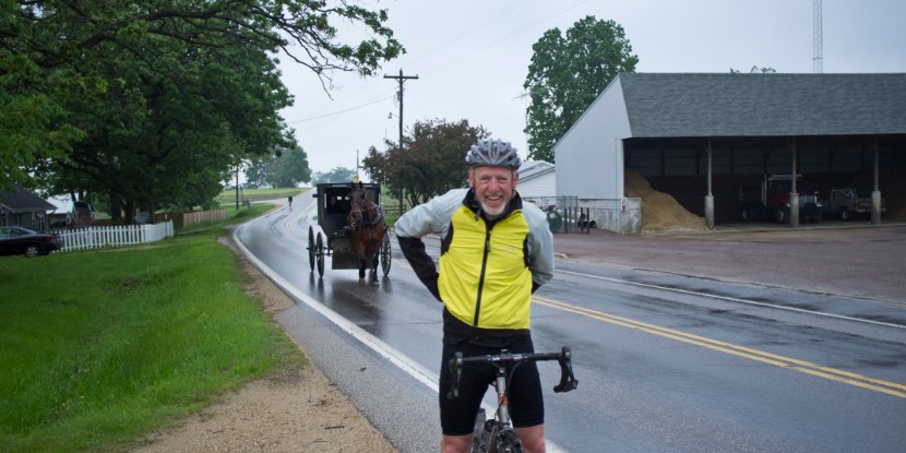 El ciclista Heinz Richardson de pie con su bicicleta mientras una carreta Amish tirada por un caballo pasa por la carretera