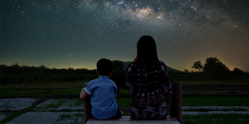 Silhouettes of a mother and child, sitting outside in nature under a starry sky.