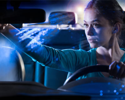 A woman driving in a car at night, adjusting the rearview mirror, with headlights behind.