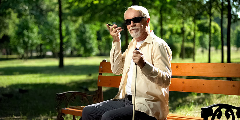 Man with vision impairment is sitting on a park bench and using a smartphone