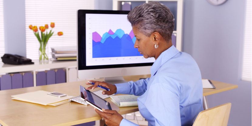 Senior businesswoman multitasking at desk with tablet device and desktop computer