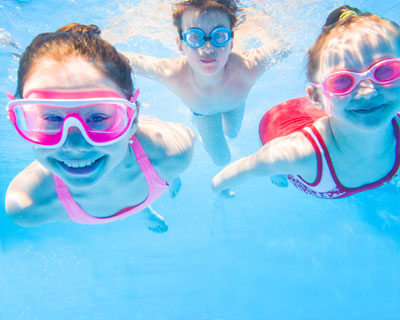 Three children swim underwater in a swimming pool, while wearing goggles.