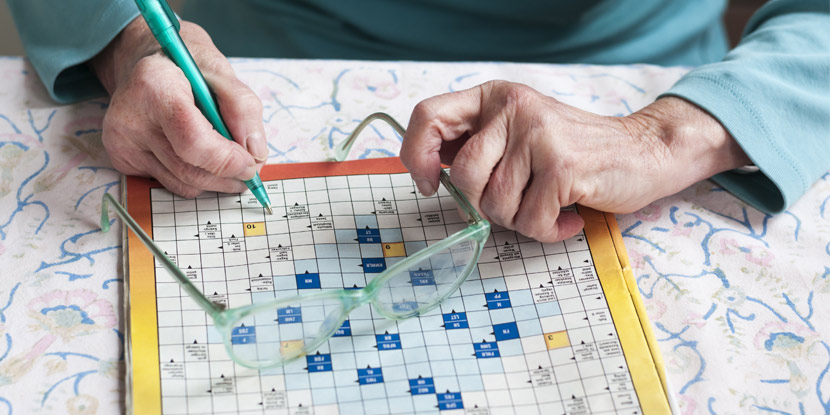 Closeup of hands of an older woman holding a pen, doing a crossword puzzle on a table with eyeglasses sitting on paper.