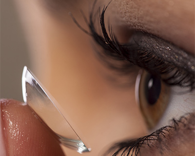 A closeup photograph of a woman putting a contact lens in her eye. The contact is on her fingertip and approaching her eye.