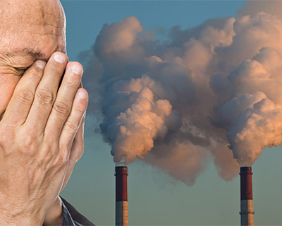 El hombre se cubre los ojos y la nariz con las manos en frente de las chimeneas con la contaminación que sale de ellos