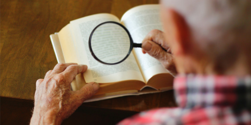 Overhead view of an older man holding a magnifying glass over a book to read.