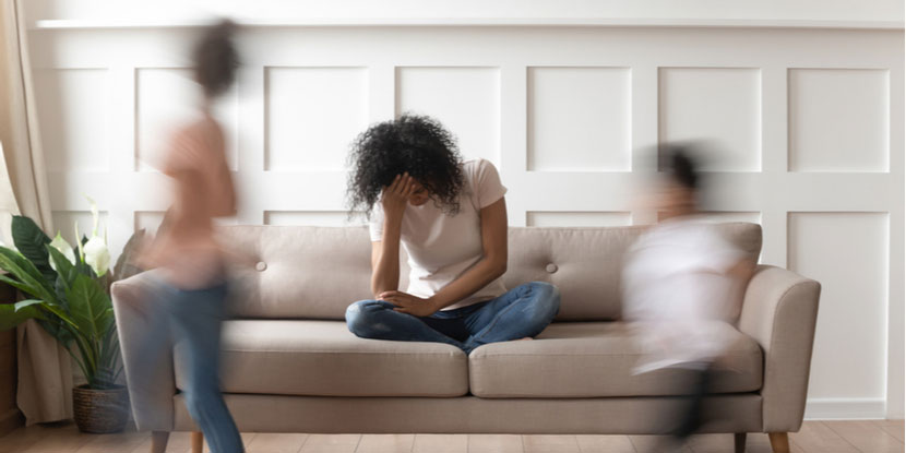 Foto de una mujer joven en su sofá sufriendo un dolor de cabeza mientras sus bulliciosos hijos corren por la casa
