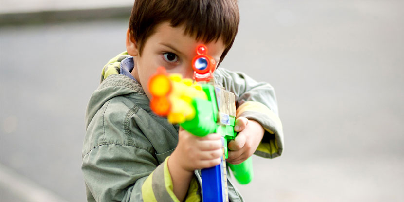 Un niño pequeño de pie, al aire libre, apunta con una pistola de juguete grande a la cámara.