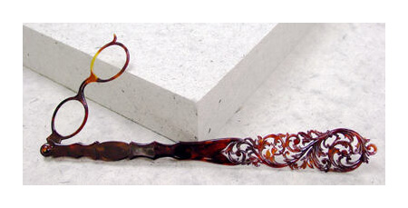 A small pair of eyeglass lenses are attached to a large carved handle. The glasses and handle are both tortoise-shell patterned, and the handle has been carved into an ornate floral pattern. The two are connected by a small hinge.