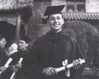 A black and white photograph of a young man holding a diploma. He is a young white man wearing a black cap and gown, and he is holding a white rolled diploma tied with a ribbon. He smiles brightly at the camera while standing in front of a tiled building.