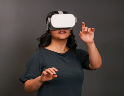 A woman wears a virtual reality headset. She is a woman with long dark hair, dark skin, and a dark blue shirt, and she wears a gray plastic virtual reality headset over her eyes. She is reaching out with one hand as if she is trying to touch something.