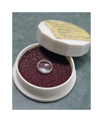 A small round clear plastic medical device sits on a dark red foam pad inside of a round white plastic case.
