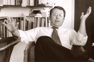 A black and white photo of a man gesturing with his hands while sitting in a chair with his legs crossed. He is a middle aged white man wearing eyeglasses, and a white shirt and dark tie. He is sitting in a wicker chair in front of a bookshelf full of books.