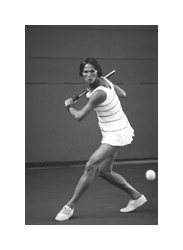 A black and white photograph of a woman playing tennis. She has short dark hair in a bob hairstyle, and she is wearing a white and black striped tennis shirt and skirt. She is holding her tennis racket behind her shoulder preparing for a backhand swing. A tennis ball can be seen coming towards her.