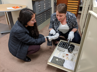 Two museum workers crouch near an open drawer full of eyeglasses. They are both women, and one has long dark hair while the other has short cropped brown hair. They are both wearing white cotton gloves and are holding up a small artifact.