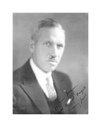 An autographed black and white photo of a man. The man is middle aged, and has gray hair and a gray toothbrush-style mustache. He wears a black suit and a patterned tie. A cursive signature is written across his left shoulder.