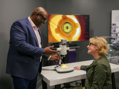 A man and a woman interact near a microscope. The man is a tall Black man in a blue suit and pink shirt, and he stands next to a white woman with shirt blonde hair and a green jacket. He is smiling and gesturing towards the microscope while she listens. Behind the microscope, there is a large television screen showing a backlit model of a human eye.