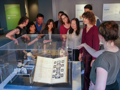A group of people listen to a museum worker while looking at a display case of old medical textbooks. The crowd is all adults, and is a mixture of men and women of different races. The museum worker is a young white woman with her hair in a bun, and she is gesturing with her hand with her back to the camera.