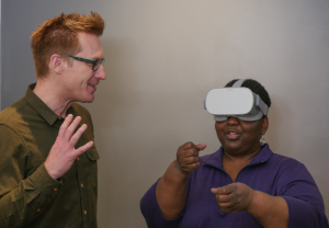 A man watches a woman use a virtual reality headset. She is a Black woman with short hair and a purple shirt, and she wears a gray plastic virtual reality headset over her eyes. He is a white man with blonde hair and eyeglass, and he turned towards her, smiling. She is gesturing with her hands like she is trying to grab something.