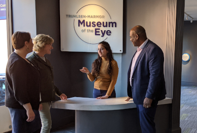 A museum volunteer interacts with three visitors at a museum front desk. The volunteer is a young woman with long dark hair wearing a yellow sweater. She gestures at two women and a man, who all small back at her. The women are both white and one has blonde hair and the other has brown hair. The man is a tall Black man with a shaved head wearing a blue suit and pink shirt. They all stand underneath a white sign with purple lettering that reads: Truhlsen-Marmor Museum of the Eye.