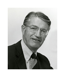 A black and white photograph of an older white man wearing a suit. He has gray, salt-and-pepper hair and he wears square framed eyeglasses.