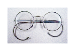 A silver pair of eyeglasses sits on a white background. The glasses have round lenses and the arms of the glasses have long hooked ends that hook behind the ear.