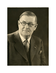 A sepia-toned photograph of a man wearing eyeglasses. He is a middle aged white man with light, slicked back hair and he wears dark round-framed glasses and a dark suit.