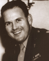 A sepia-toned photograph of a smiling man in a military uniform. He is a middle aged white man with dark hair, and he wears a dark Army uniform.