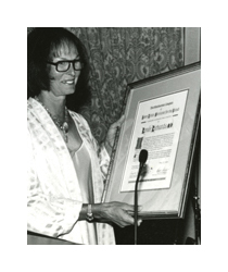 A black and white photograph of a woman holding a framed award. She has short dark hair in a bob hairstyle, and she wears dark rimmed eyeglasses and a long white sweater. She is holding a large wooden frame with an award certificate mounted inside. There is a long, thin black microphone in front of her.