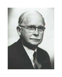 A black and white photograph of an older white man. He has thin, white hair and a white mustache. He is wearing eyeglasses and a dark suit.