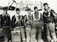 A black and white photograph of five young men in pilot's uniforms standing in front of an airplane cockpit. They are all young men wearing leather bomber jackets and dark hats, and they all have white parachute straps across their chest. The man on the far left is wearing dark, aviator-style sunglasses.